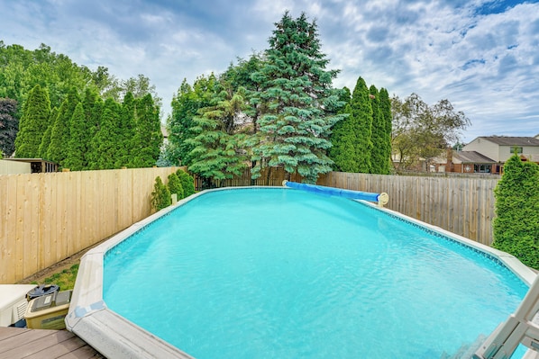 A rectangular swimming pool with a wooden deck surrounding it, set against a backdrop of a wooden fence and lush green trees.