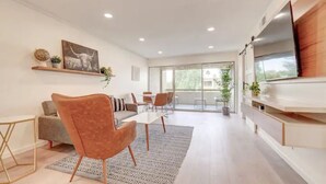 A spacious living area with a sofa, a coffee table, and a dining table with chairs. There is a large window providing natural light and a view of greenery outside. - Walk to Old Town. Heated Pool. Hot tub. Gym. (Scottsdale)