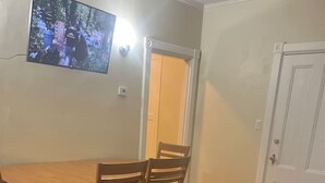 Un salon comprenant une table à manger en bois, deux chaises en bois, un canapé en cuir noir et un téléviseur fixé au mur.