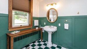 Un baño con tocador de madera, lavamanos pedestal, un espejo grande con marco dorado y una ventana con persianas.