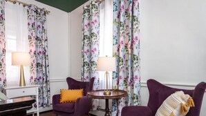 Una habitación con sillones morados, una pequeña mesa redonda con lámparas y cortinas con estampado floral.