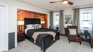 Une chambre avec un mur d’accent en briques, un lit avec une tête de lit sombre, deux tables de chevet avec des lampes, un fauteuil en cuir marron et une fenêtre avec des rideaux.