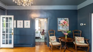 A room with dark blue walls, wooden flooring, and a chandelier. There are two wooden chairs with cushions, a round table with a vase of flowers, and framed pictures on the wall.