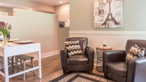 A modern living room with a white dining table, two leather armchairs, a small round table with a bottle, and a painting of the Eiffel Tower.