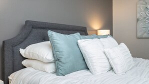 A neatly made bed with a grey headboard, white and teal pillows, and a wall-mounted lamp. - The Sugar Maple - Suite (Harrisonburg)