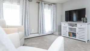 A modern living room with a large flat-screen TV, a white entertainment unit, and light-colored furniture. - The Sugar Maple - Suite (Harrisonburg)