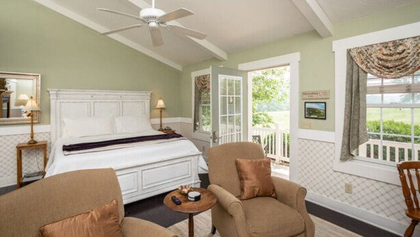 Une chambre à coucher comprenant un lit, deux chaises, une petite table, un ventilateur de plafond et une fenêtre donnant sur un paysage verdoyant.