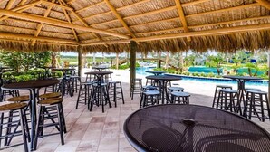 Un espace extérieur aménagé avec des tables et des chaises, abrité par un toit de chaume, et offrant une vue sur une piscine et la verdure environnante.