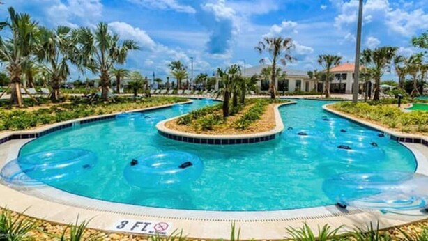 Une grande piscine extérieure dotée de plusieurs couloirs, entourée de palmiers et surplombée d’un ciel bleu azur.
