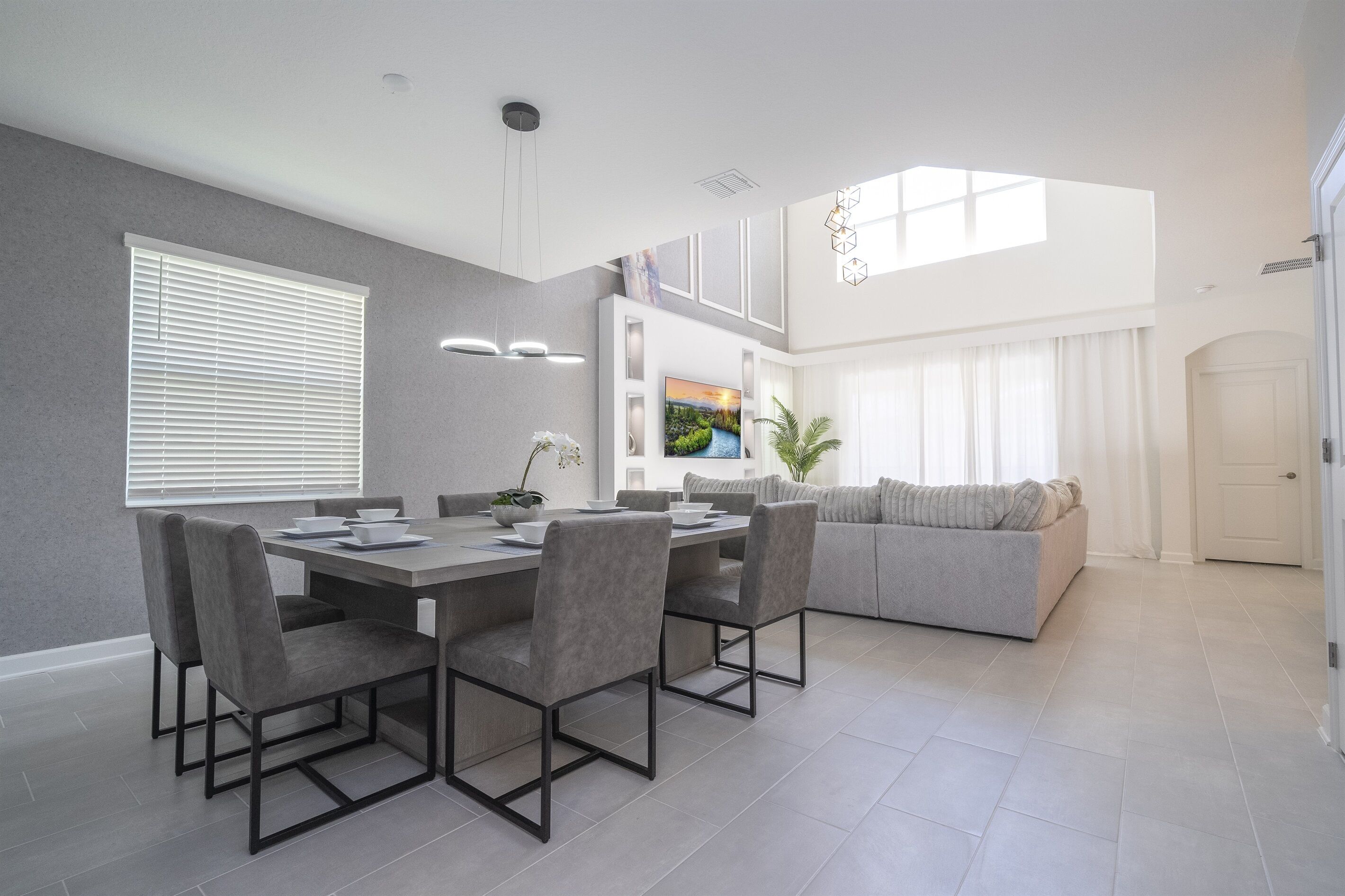 Un espace repas moderne avec une table grise et des chaises assorties, un canapé gris et une grande fenêtre avec des stores.