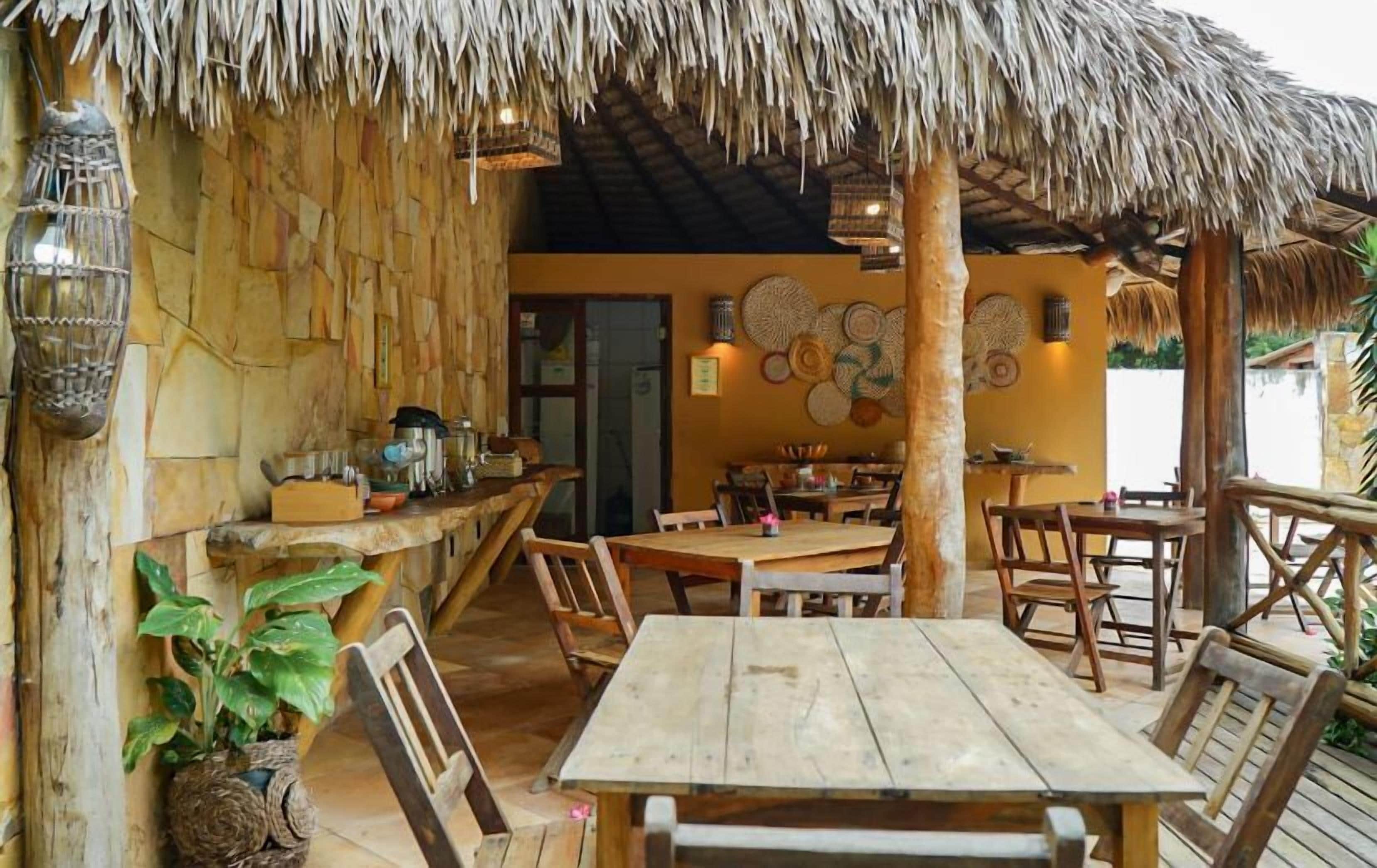 Un espacio de comedor al aire libre con mesas y sillas de madera, techo de paja y decoraciones murales tejidas.