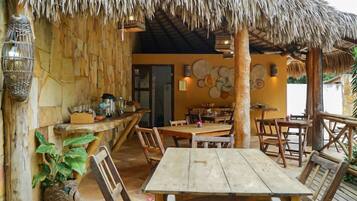 Un espacio de comedor al aire libre con mesas y sillas de madera, techo de paja y decoraciones murales tejidas.