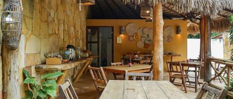 An outdoor dining area with wooden tables and chairs, a thatched roof, and woven wall decorations.