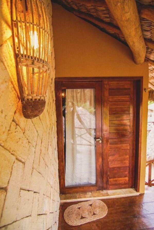 A wooden door with a glass panel, a woven wall hanging, and a stone wall. - Pousada Rota dos Ventos (Cajueiro da Praia)