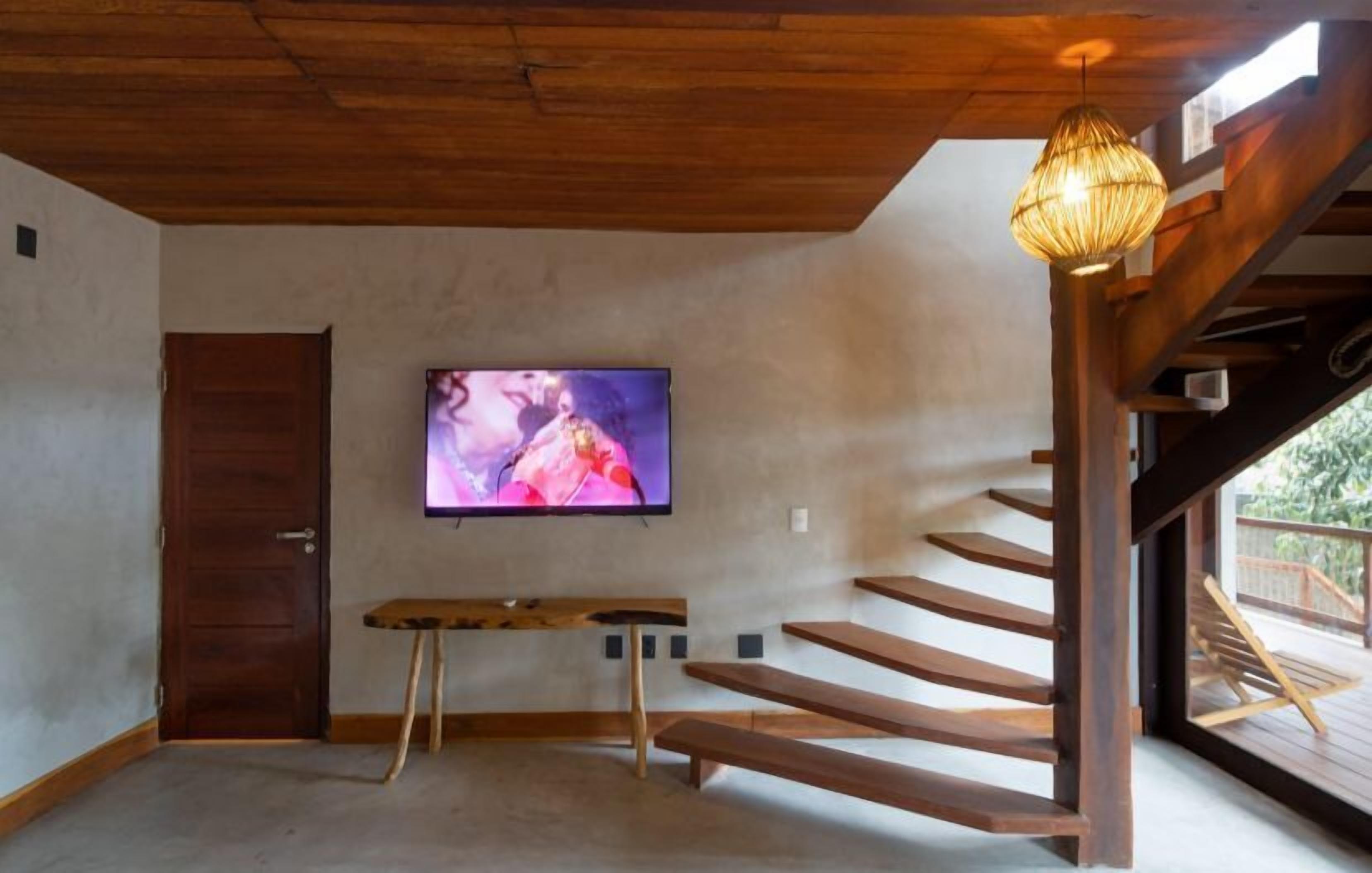 Una habitación con una escalera de madera, un televisor instalado en la pared y un pequeño taburete de madera.