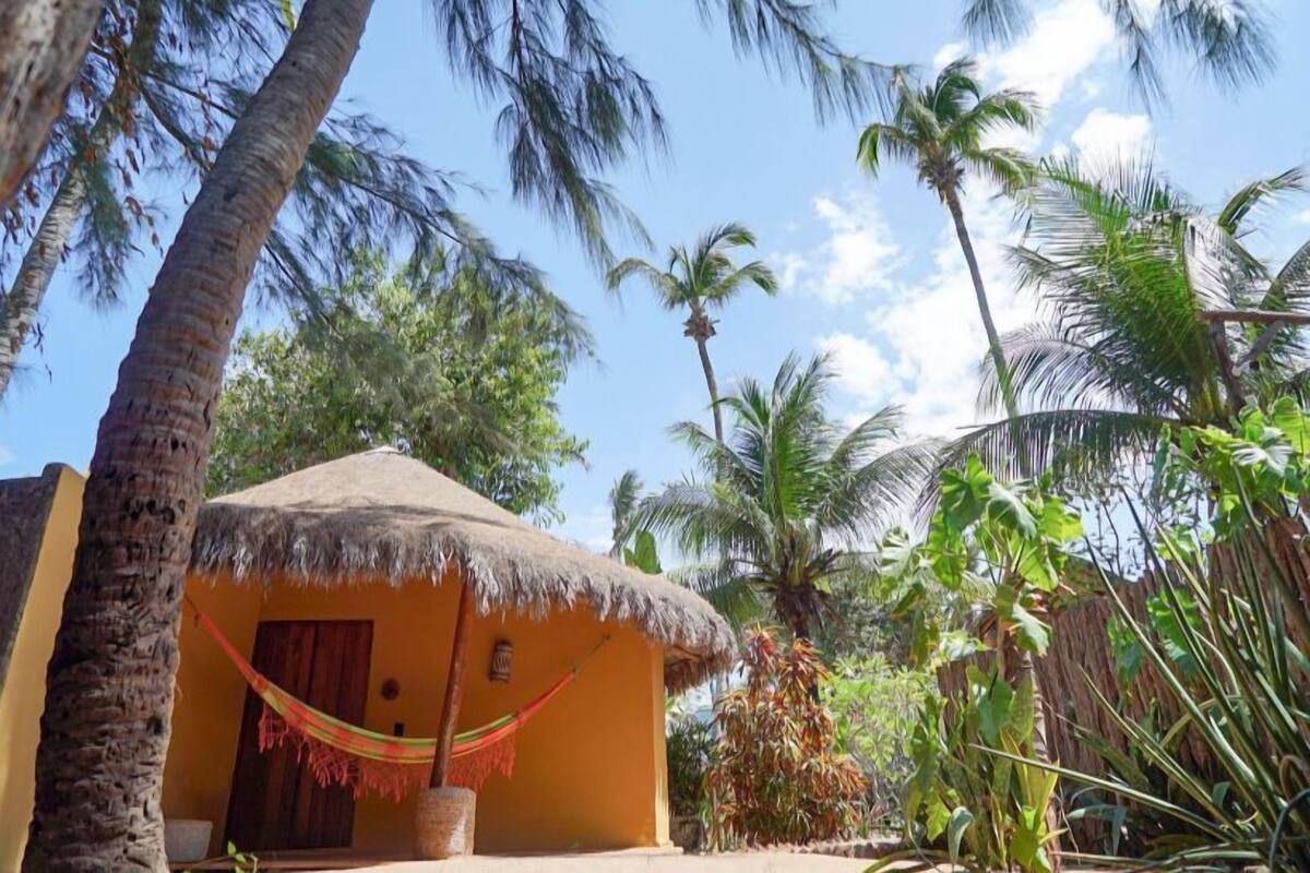 A thatched-roof hut with a hammock, surrounded by palm trees and tropical vegetation.