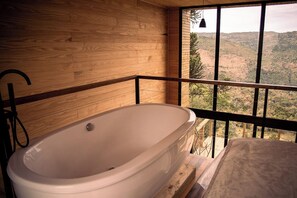 A modern bathroom with a freestanding bathtub, wooden walls, and a balcony with a view of a hilly landscape.