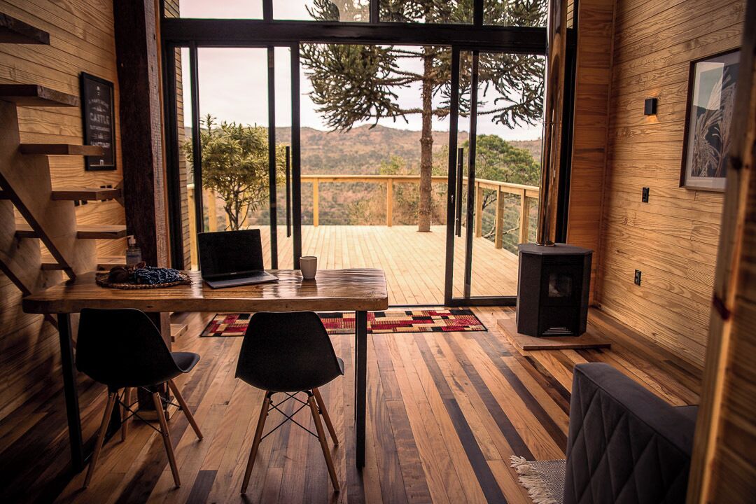 A modern interior with a wooden floor, a dining area with a laptop and chairs, a fireplace, and a large window offering a view of trees and hills.