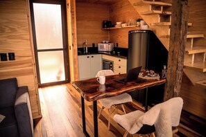 A compact kitchen with a microwave, toaster, and refrigerator. There is a wooden table with a laptop and a mug. A sofa and a chair are visible in the foreground.