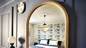 A bedroom with a large mirror, a bed with a patterned headboard, a nightstand with a lamp, and a wall-mounted clock. - Maison D'hôte - Chambre Double Kaolin (LIMOGES)