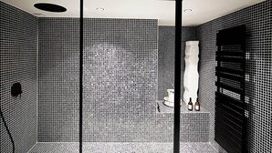 A bathroom with a tiled floor and walls, a shower area with a black door, and a white towel rack with bottles. - Maison D'hôte - Chambre Double Kaolin (LIMOGES)