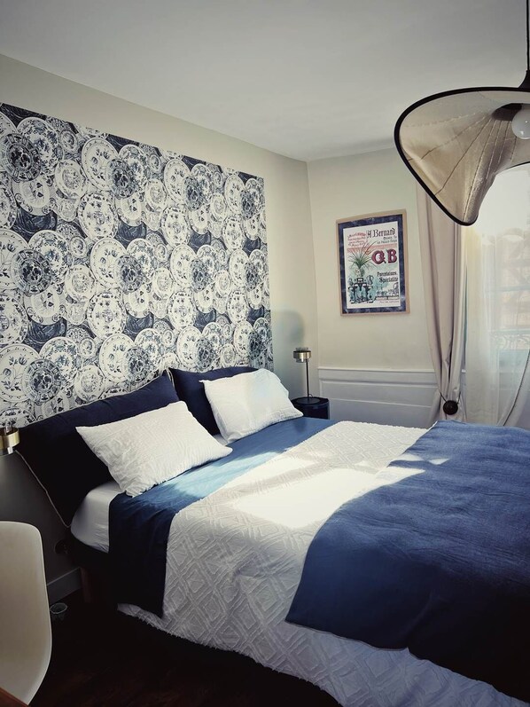 Une chambre avec un papier peint à motifs, un lit avec une literie bleue et blanche, une table de chevet, une lampe et un tableau encadré au mur.