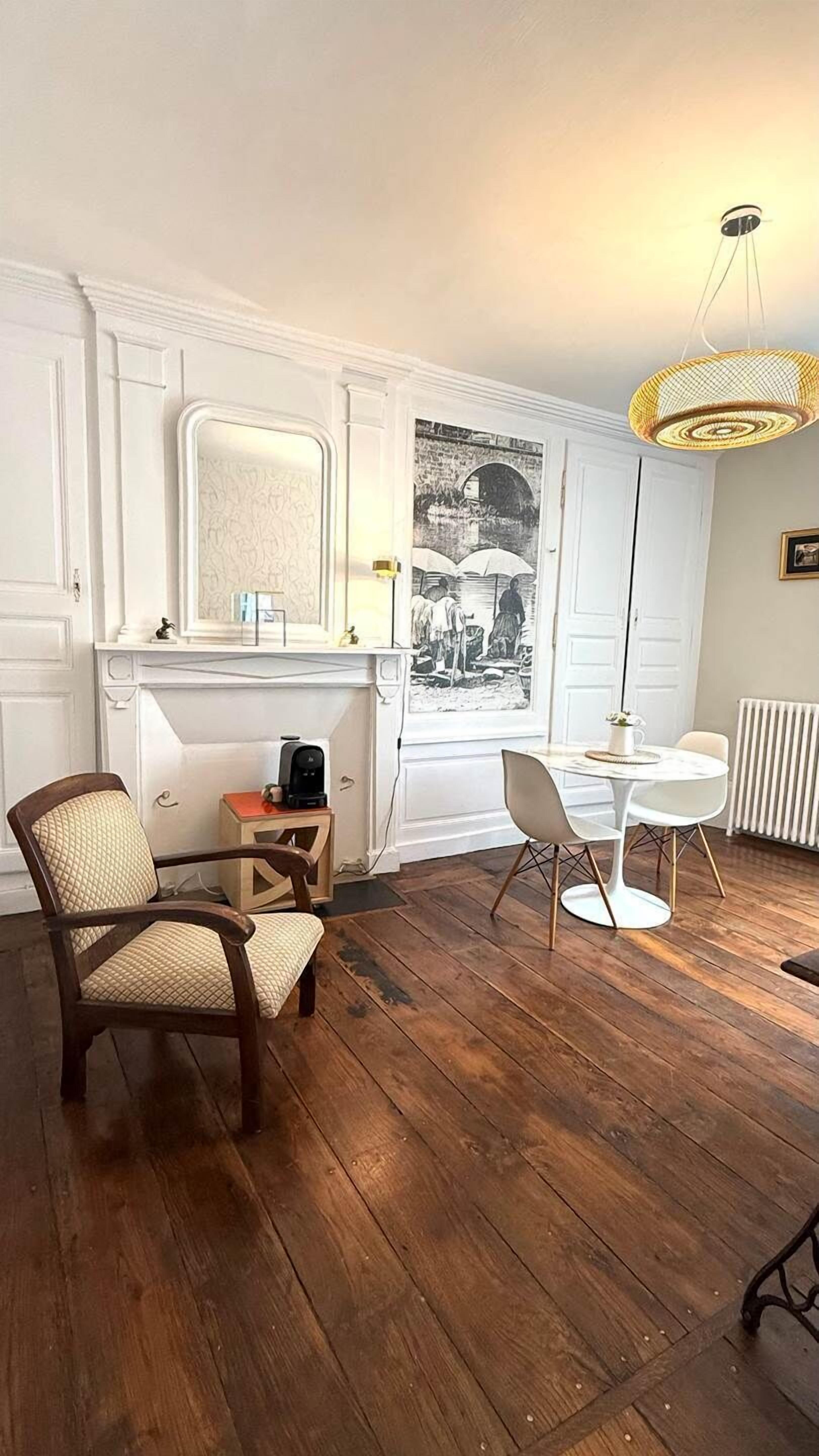 A room with a wooden floor, white walls, a white door, a white table with two chairs, a wooden chair, a small table with a kettle, and a hanging light fixture.