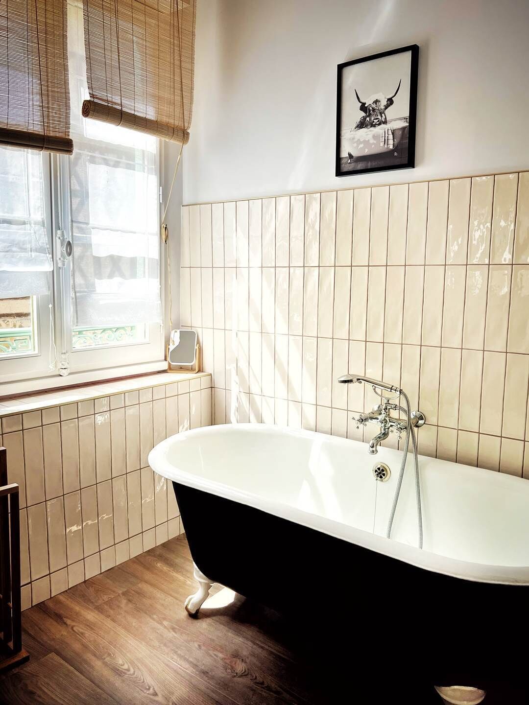 Une salle de bain avec une baignoire sur pieds, une fenêtre avec des stores et un tableau encadré au mur.