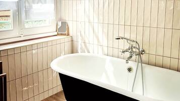 Une salle de bain avec une baignoire sur pieds, une fenêtre avec des stores et un tableau encadré au mur.