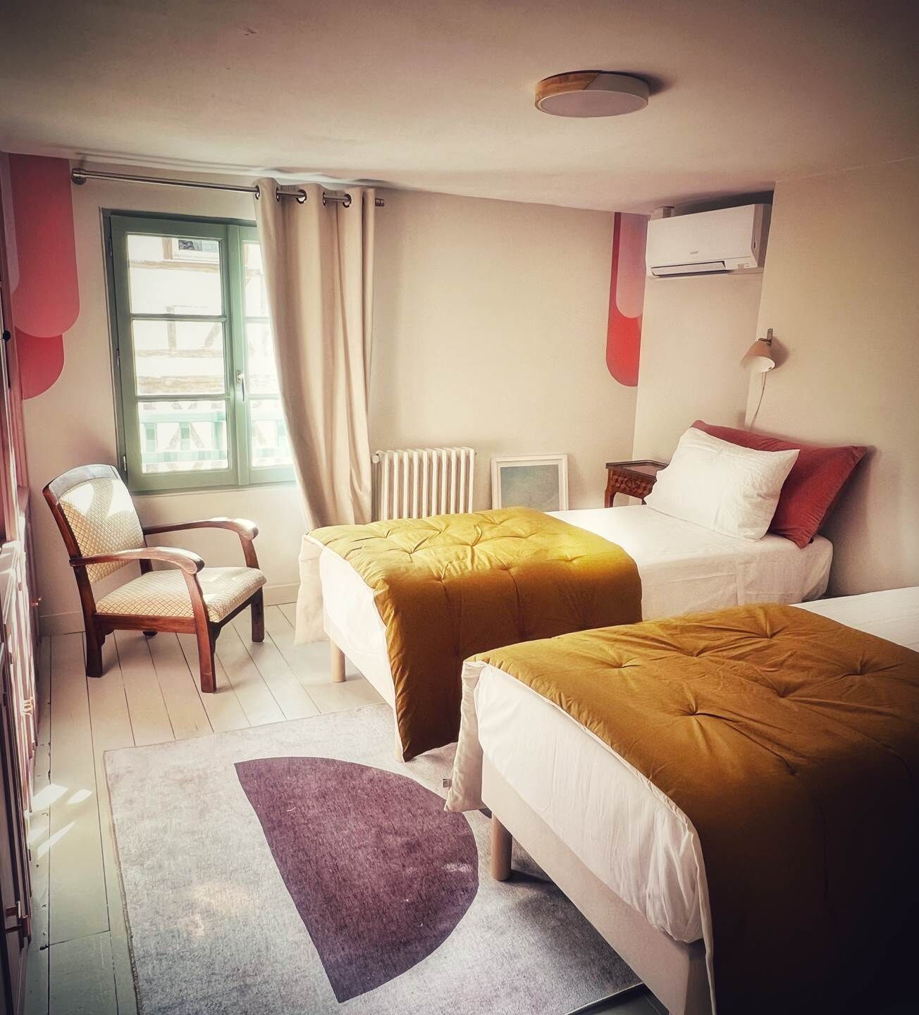 Quarto de hotel com duas camas, uma cadeira, um radiador, uma janela com cortinas e uma luminária fixada na parede.