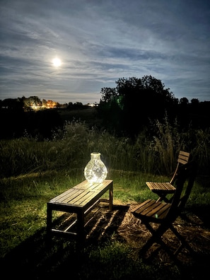 Un banc et une table en bois, avec un vase en verre illuminé, dans un décor extérieur verdoyant, la nuit.