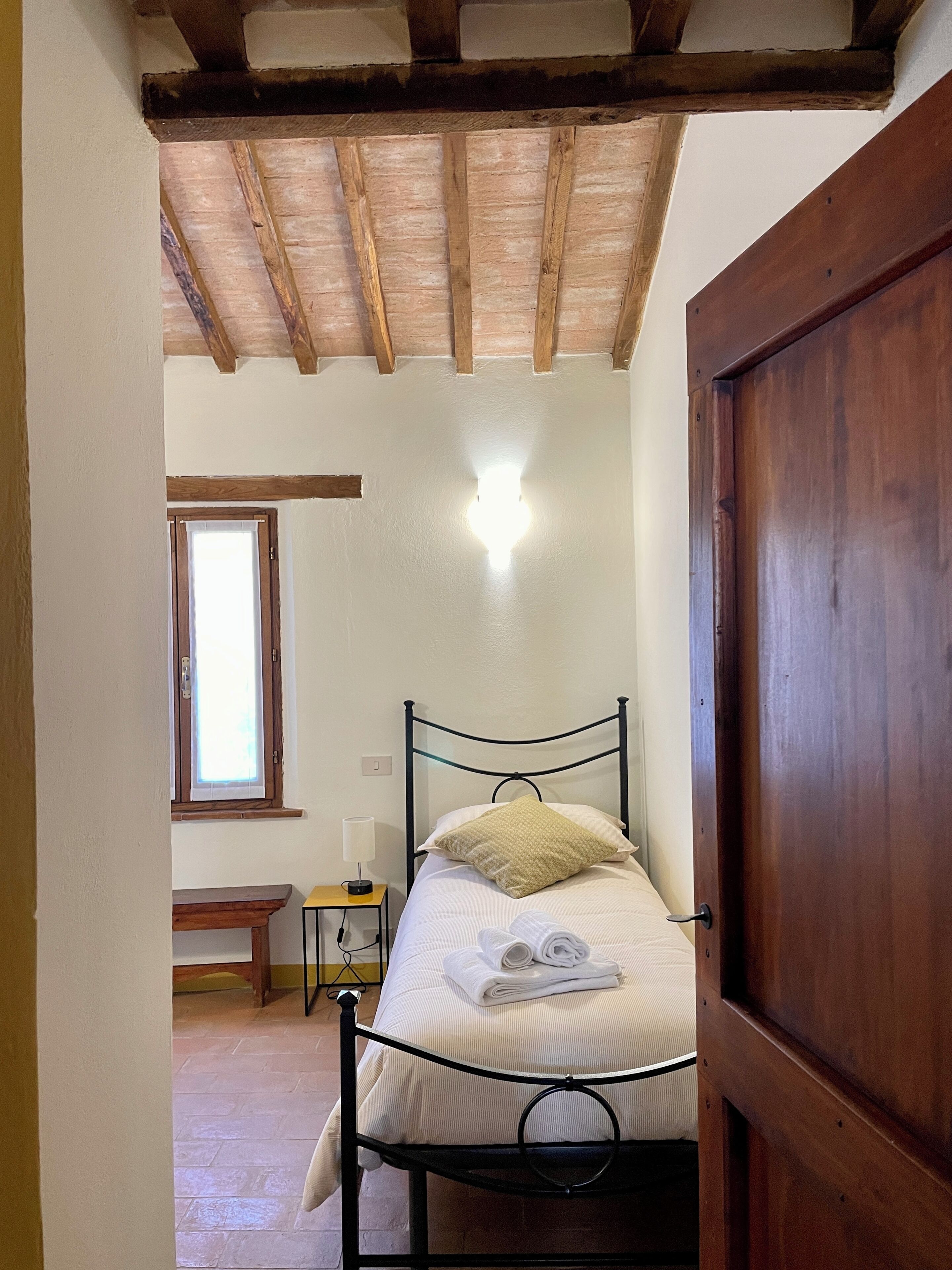 Um quarto com uma cama, uma mesa de cabeceira, uma luminária, uma janela e uma porta de madeira.