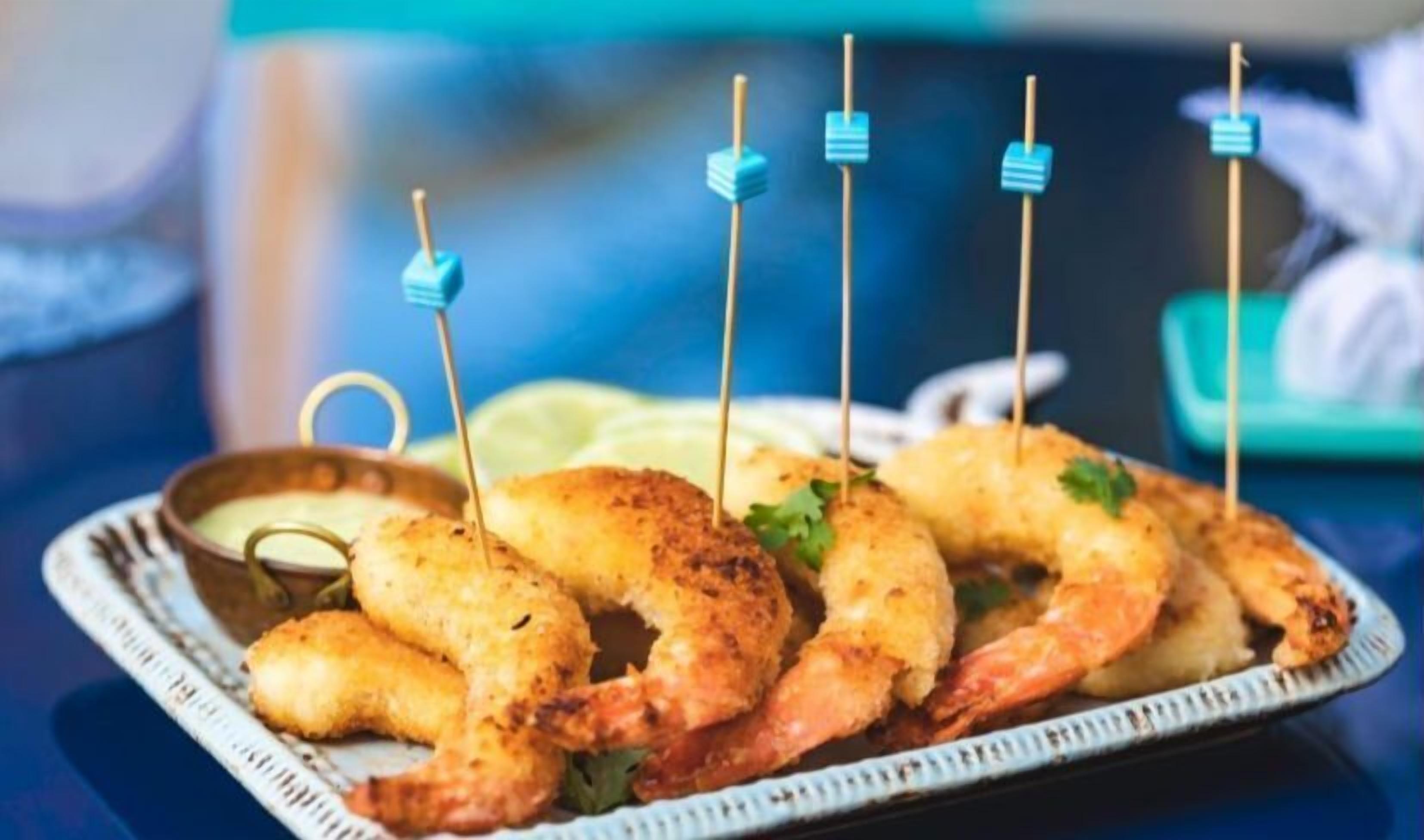 A plate of shrimp skewers with toothpicks, served with a side of sauce and lemon wedges.