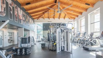 A gym with various exercise equipment, including treadmills and weight machines, under a wooden ceiling with large windows.
