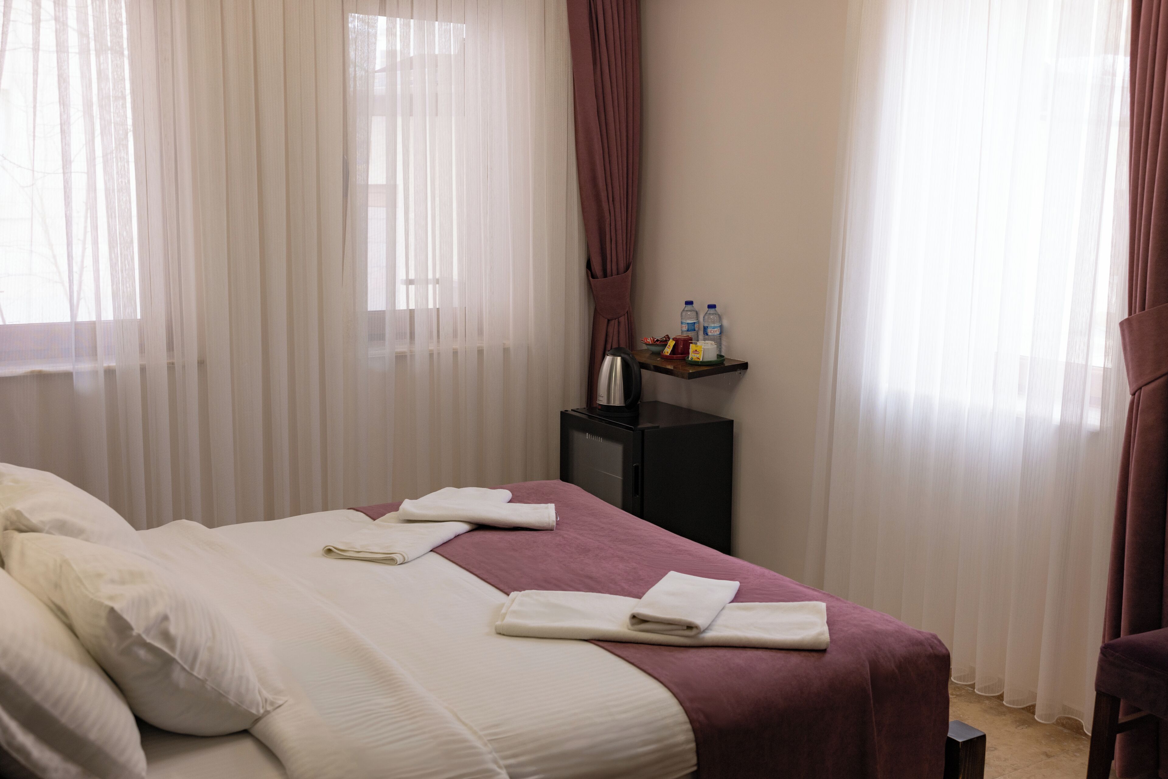 Quarto de hotel com cama, almofadas, mesa de cabeceira com abajur e janela com cortinas.