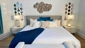 A large bed with a white headboard, blue and white pillows, and a blue throw blanket. There are two bedside tables with lamps and decorative wall art above the bed. - Verde Vista | Steam Shower | Theatre | Hot Tub | Games (East Kootenay)
