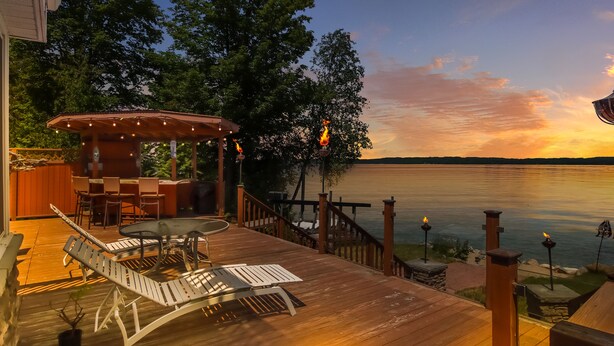 Une terrasse en bois avec des meubles d’extérieur, un espace salon couvert et une vue sur un lac au coucher du soleil.
