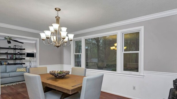 A dining area with a wooden table and four chairs, a chandelier, and large windows with a view of trees. - The Quail's Nest of Harbison | Sleeps 8 (Columbia)