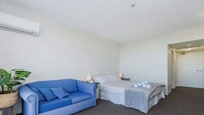 A room with a blue sofa, a bed with white bedding, a bedside table with a lamp, and a potted plant. - Studio 211-seaside Serenity: Your Perfect Retreat (North Ward)