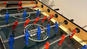 Een tafelvoetbaltafel in een kamer met een aan de muur bevestigde televisie en een plant in de hoek.