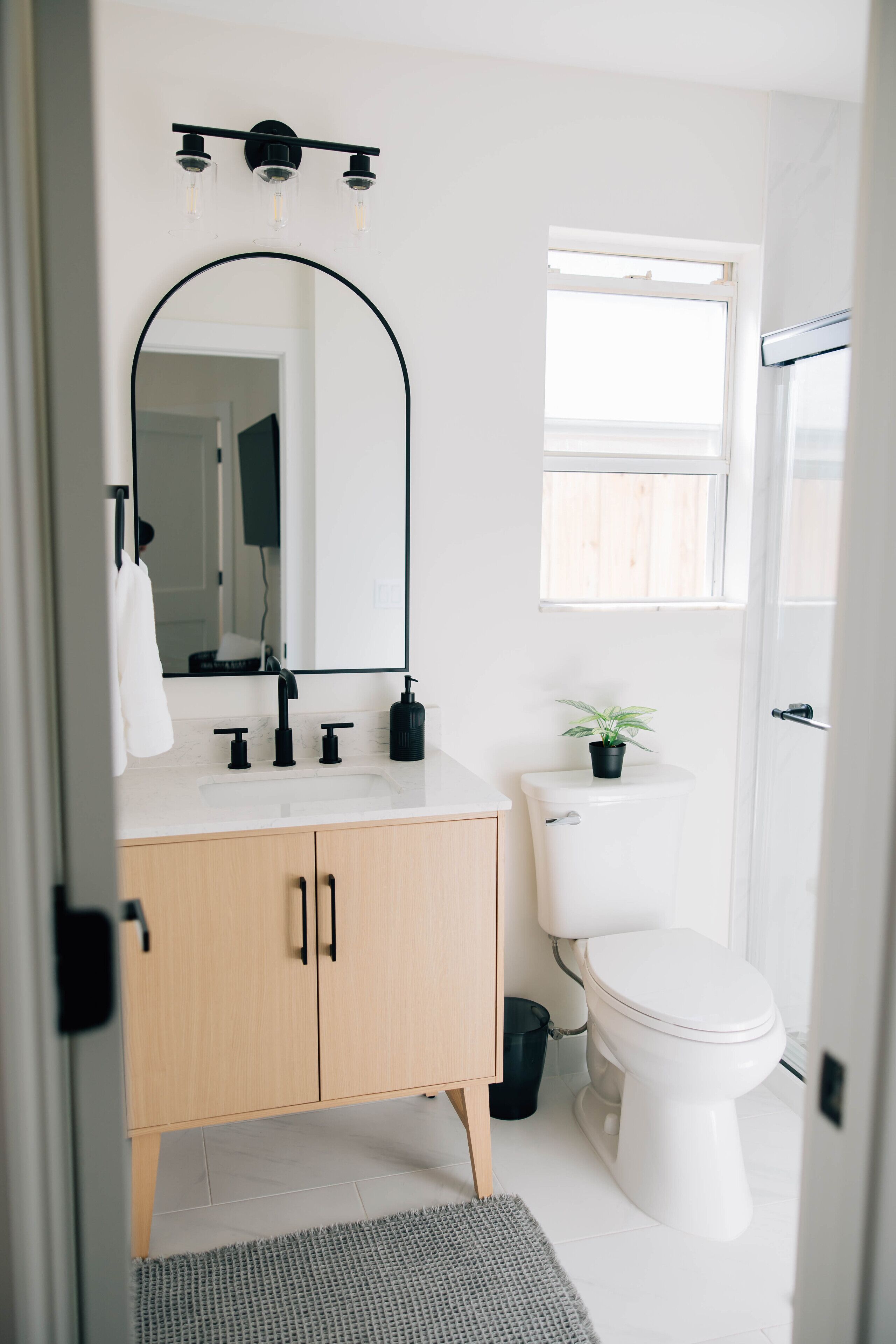 A modern bathroom with a white sink, black faucet, and a large mirror above it. There is a toilet with a potted plant beside it, and a window above the toilet.