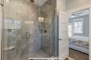 A modern bathroom with a glass-enclosed shower, a white door, and a visible bedroom with a bed and a star-patterned rug. -  Newer OBX home just 350 yards to the beach. Private Pool & Hot Tub. (Corolla)
