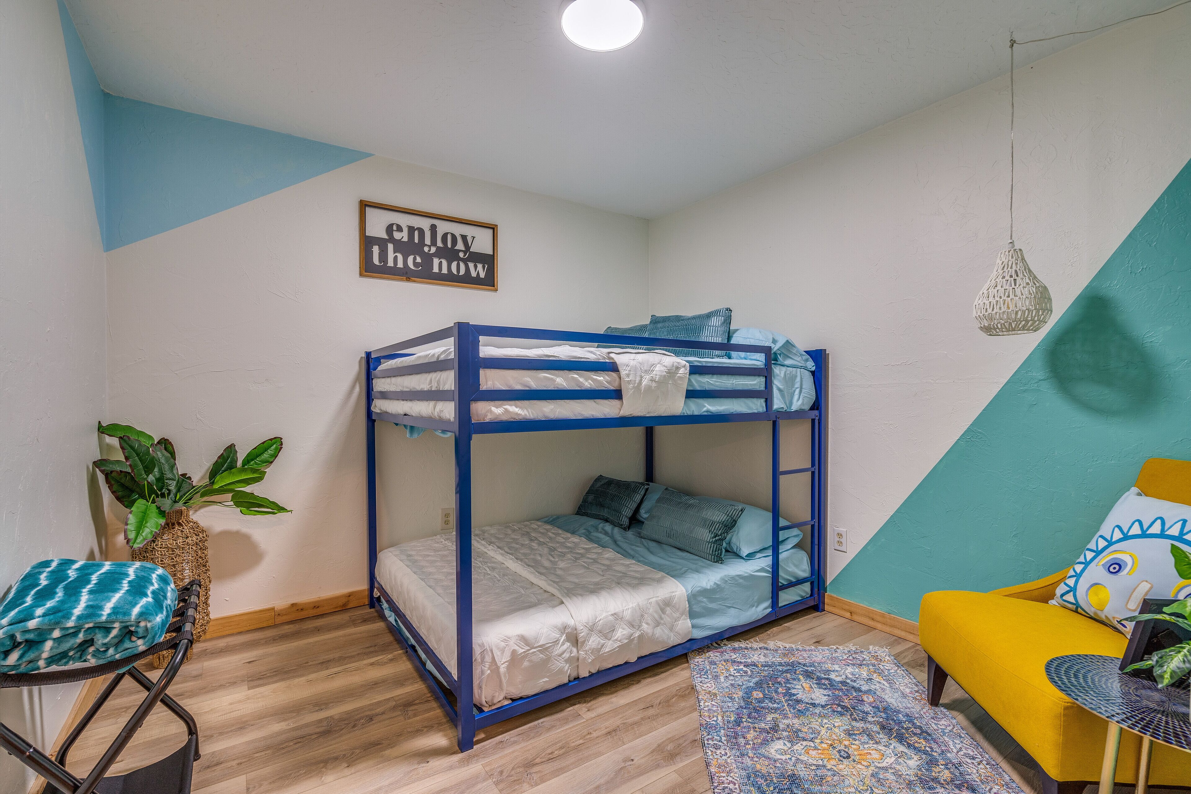 Una cama litera con estructura azul, una mesita con una planta, un sillón amarillo y una alfombra estampada.