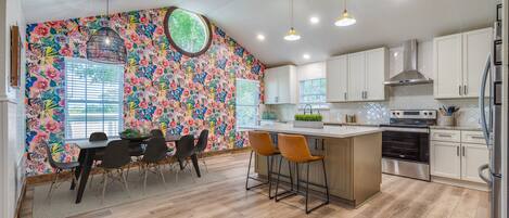 A kitchen with wooden flooring, white cabinets, a central island with stools, and a colorful floral wall.