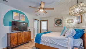 Ein Schlafzimmer mit einem Bett, einem Fernseher auf einer Holzkommode, einem Deckenventilator und Kunstwerken an den Wänden.