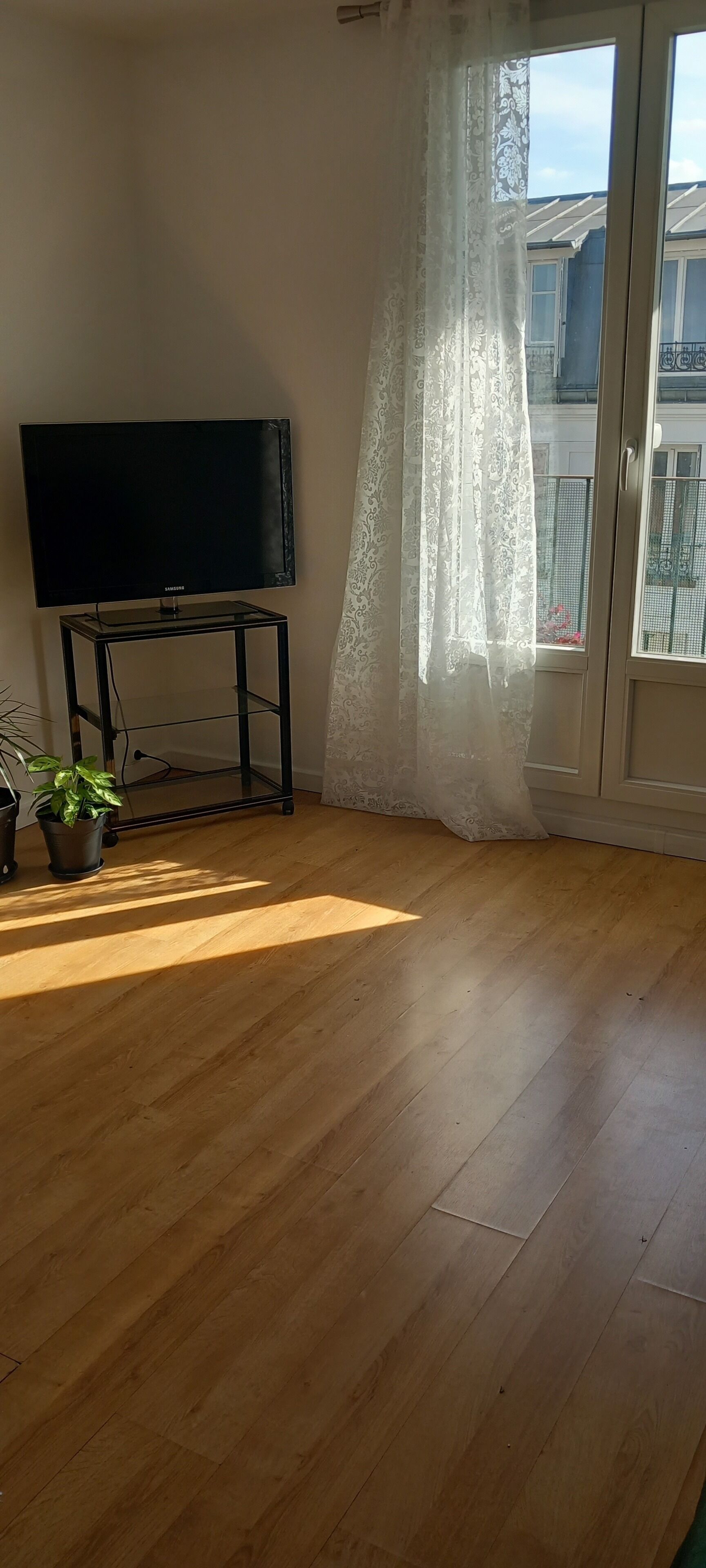 A room with a wooden floor, a black TV on a glass stand, and a window with a white curtain.