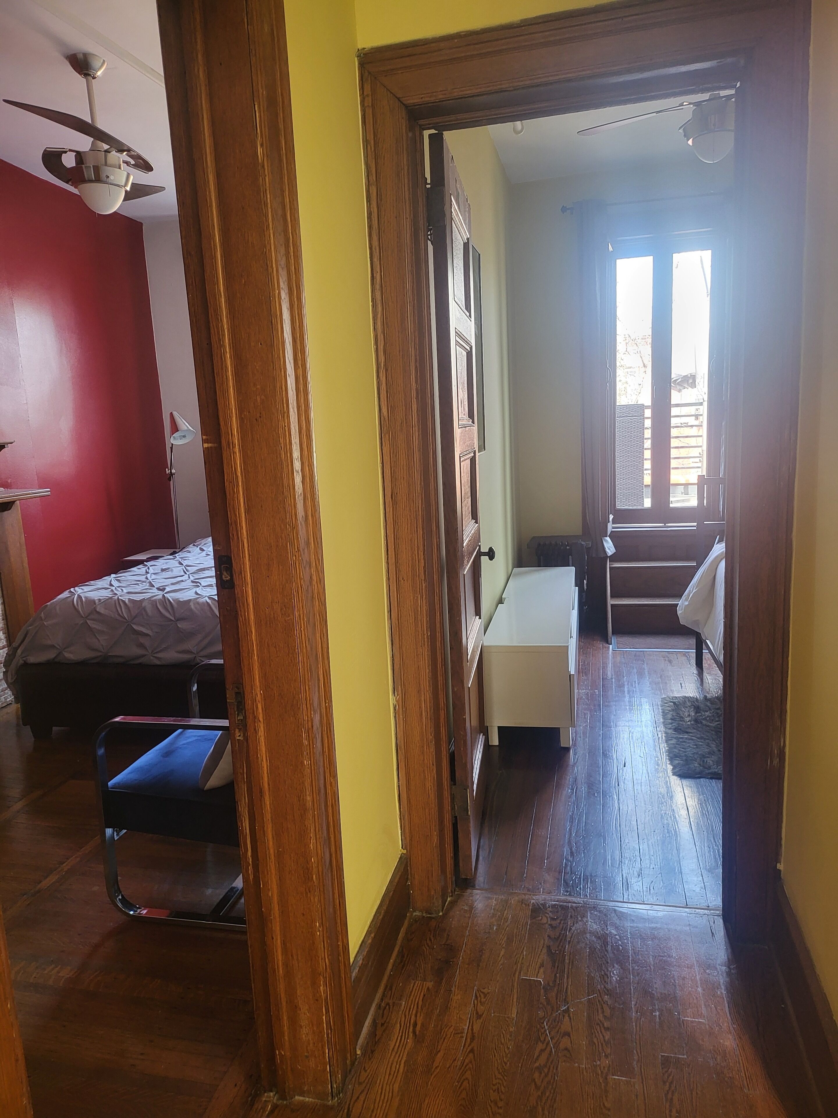 A hallway with wooden flooring, a bed with a red wall, a desk, and a window with a view of the outside.