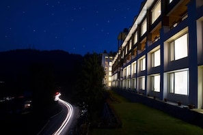 Un hotel de varios pisos con balcones y ventanas amplias, un camino curvo con estelas de luz, y un cielo nocturno estrellado.
