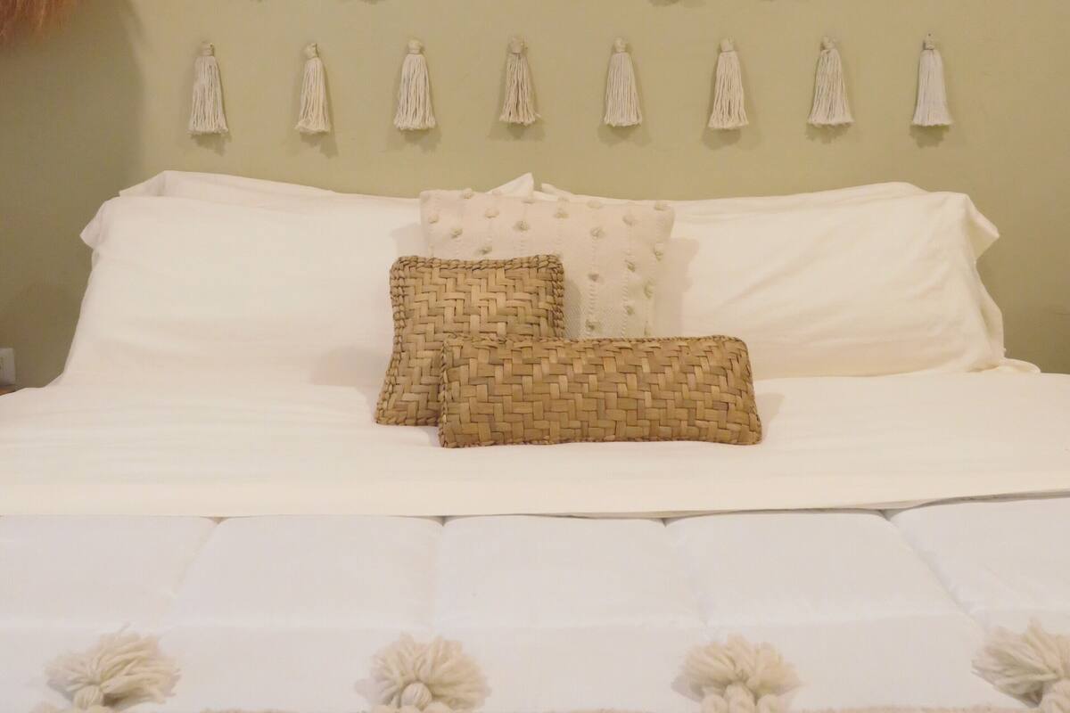 A bed with white bedding, two woven pillows, and a wall with hanging tassels.