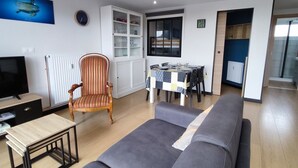 A living room with a sofa, a TV, a wooden chair, a dining table with a patterned tablecloth, and a cabinet with glass doors. - Appartement 4 Personnes à Berck (Berck-sur-Mer)